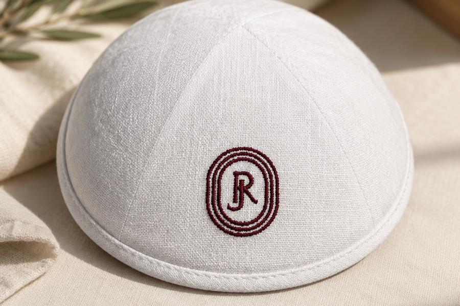Kippa blanche en lin personnalisée avec un monogramme bordeaux brodé, photographiée dans une ambiance lumineuse et raffinée.
