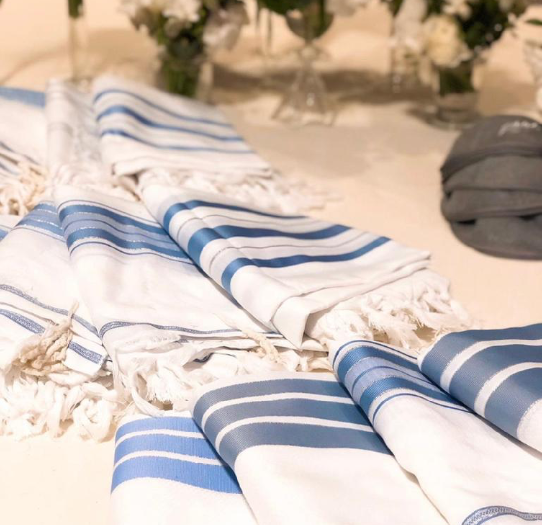 Talit blanc personnalisé avec rayures bleues et franges traditionnelles, présenté plié sur une table lors d'une cérémonie.
