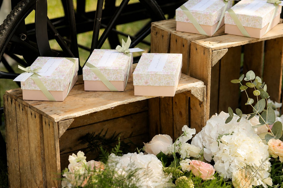 Coffrets cadeaux personnalisés à motif floral avec rubans en satin vert sauge, présentés sur des caisses en bois lors d'une décoration de mariage élégante.