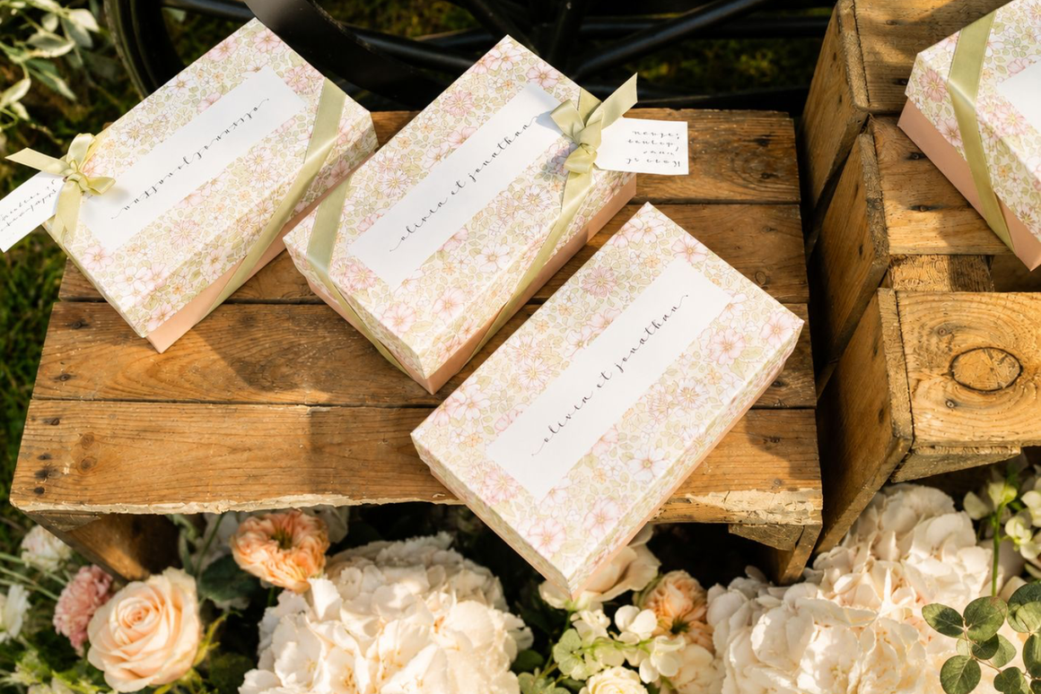 Coffrets cadeaux personnalisés à motif floral avec rubans en satin vert sauge, présentés sur des caisses en bois lors d'une décoration de mariage élégante.