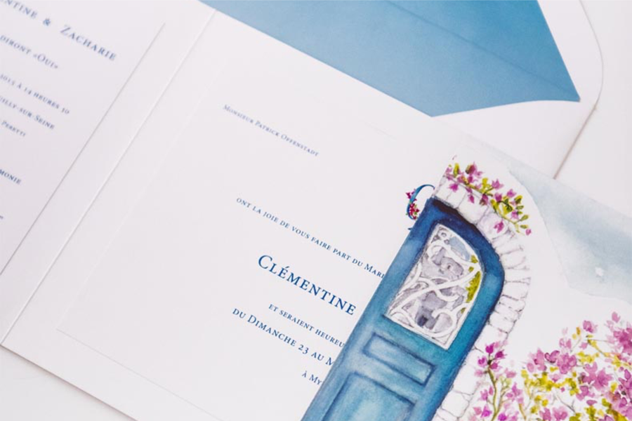 Faire-part de mariage personnalisé Mykonos signé BL Creations, illustré d'une porte bleue typique des Cyclades et de bougainvilliers en fleurs. Présenté avec une enveloppe blanche doublée de bleu, ce modèle évoque l'élégance, le charme méditerranéen et l'atmosphère romantique de l'île de Mykonos.