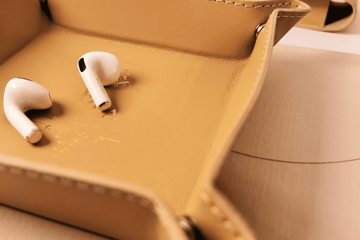 Plateau vide-poche personnalisé en cuir avec monogramme doré, contenant une paire d'écouteurs sans fil, idéal comme cadeau personnalisé ou accessoire de bureau élégant.